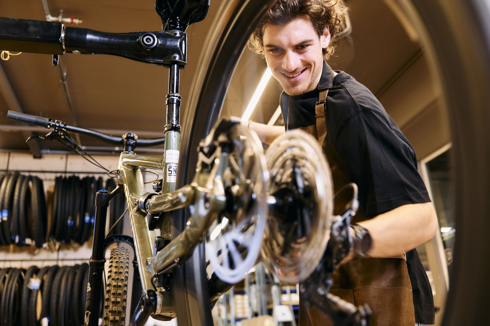 Eine Person, die eine Schürze trägt, lächelt, während sie in einer Werkstatt an einem Fahrrad arbeitet. Sie justiert das Hinterrad, im Hintergrund sind Regale mit Reifen zu sehen.