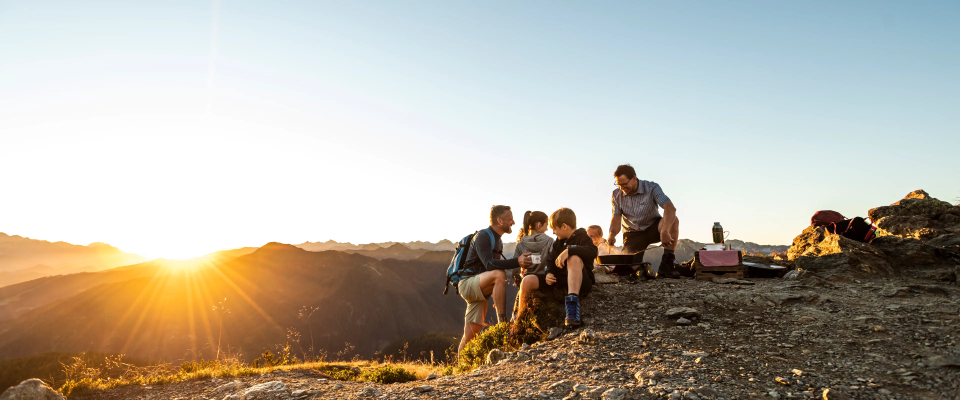 Eine Gruppe von Menschen sitzt bei Sonnenuntergang auf einem felsigen Hügel, im Hintergrund sind Berge zu sehen und die Sonne wirft einen warmen Schein über die Landschaft.
