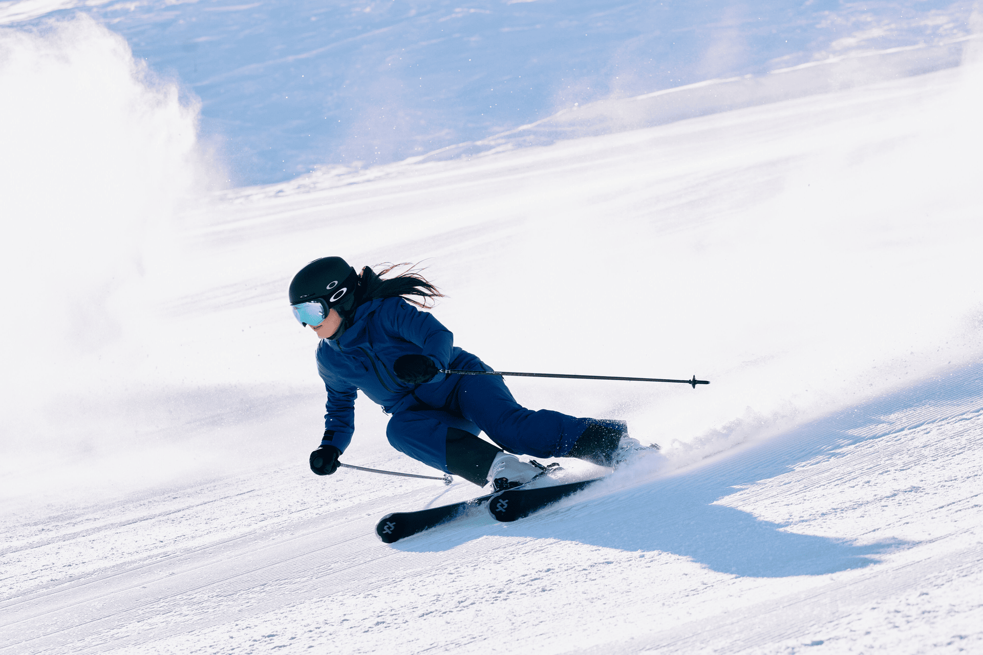 Ein Skifahrer in blauem Outfit und Helm fährt eine scharfe Kurve auf einem verschneiten Hang und erzeugt dabei eine Schneespritze hinter sich. Die Sonne wirft einen Schatten auf den Schnee und unterstreicht die dynamische Bewegung des Skifahrers.