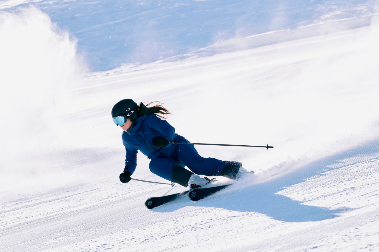 Ein Skifahrer in blauem Outfit und Helm fährt eine scharfe Kurve auf einem verschneiten Hang und erzeugt dabei eine Schneespritze hinter sich. Die Sonne wirft einen Schatten auf den Schnee und unterstreicht die dynamische Bewegung des Skifahrers.