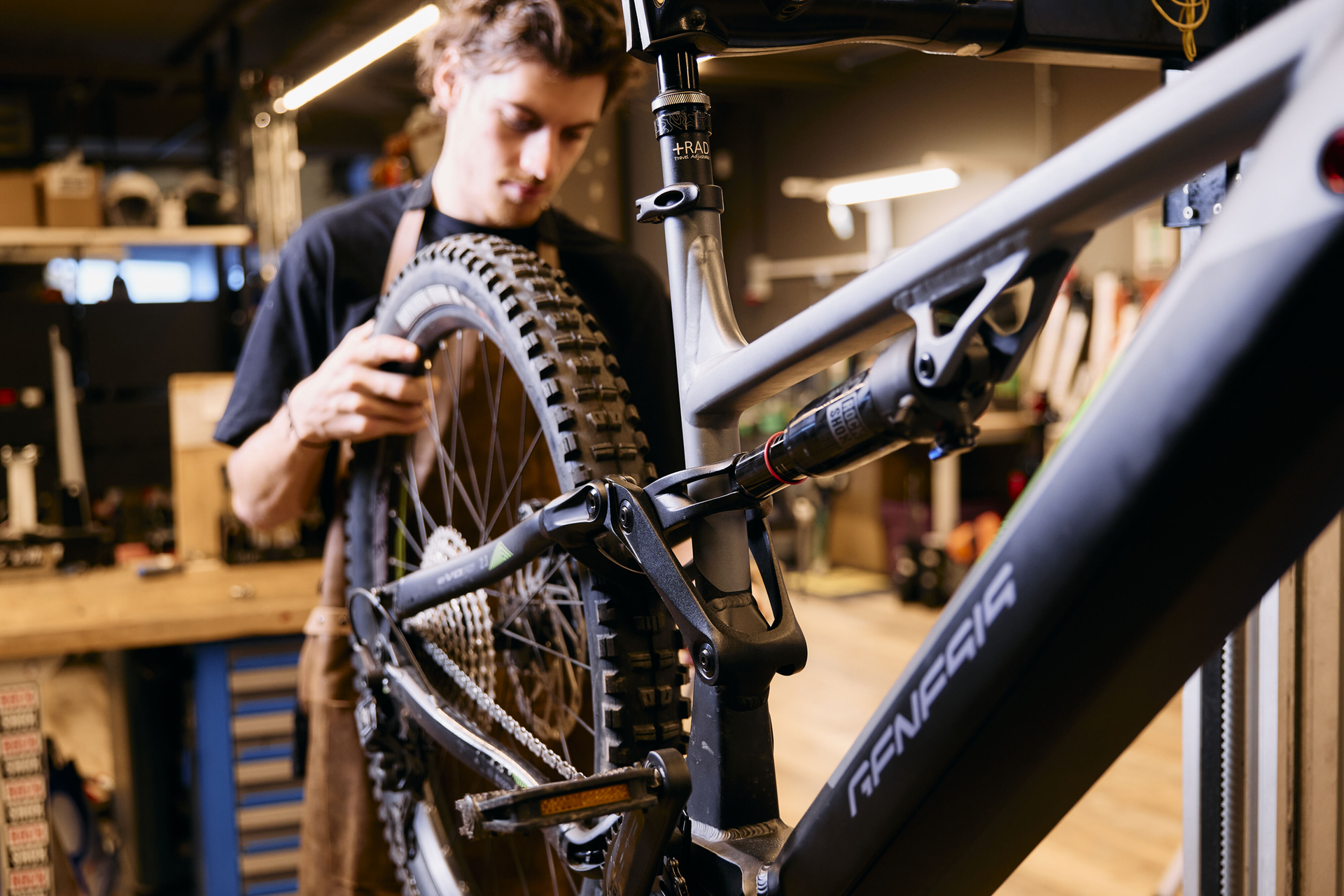 Eine Person arbeitet in einer Werkstatt an einem Mountainbike und konzentriert sich dabei auf das Hinterrad und die Federung. Die Werkstatt ist mit Werkzeugen ausgestattet, im Hintergrund steht eine Werkbank.