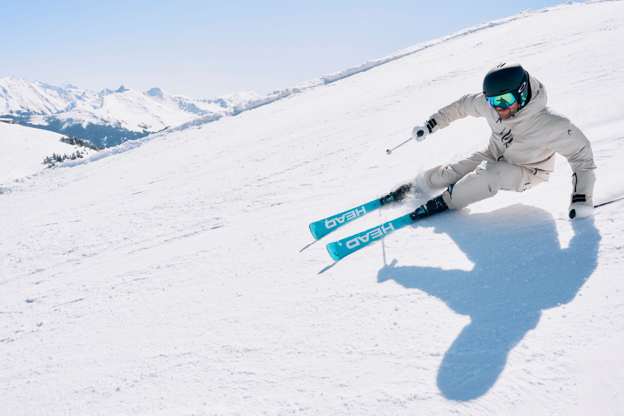 Ein Skifahrer in weißem Outfit und Helm fährt eine scharfe Kurve auf einem verschneiten Hang, im Hintergrund blauer Himmel und ferne Berge.