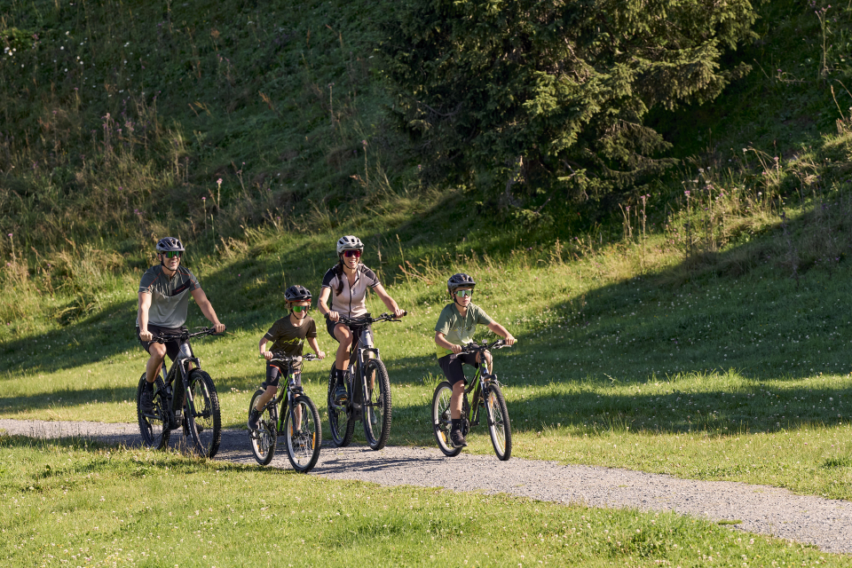Eine vierköpfige Familie mit Helmen radelt auf einem Schotterweg durch eine Grasfläche mit Bäumen im Hintergrund. Die Gruppe besteht aus zwei Erwachsenen und zwei Kindern, alle fahren Mountainbikes.