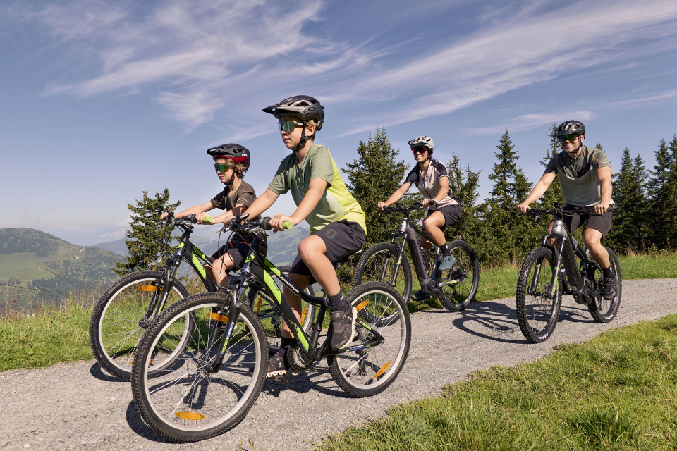Vier Personen mit Helmen fahren mit dem Fahrrad auf einem Schotterweg durch eine malerische Landschaft mit grünem Gras und Bäumen unter einem blauen Himmel mit dünnen Wolken.