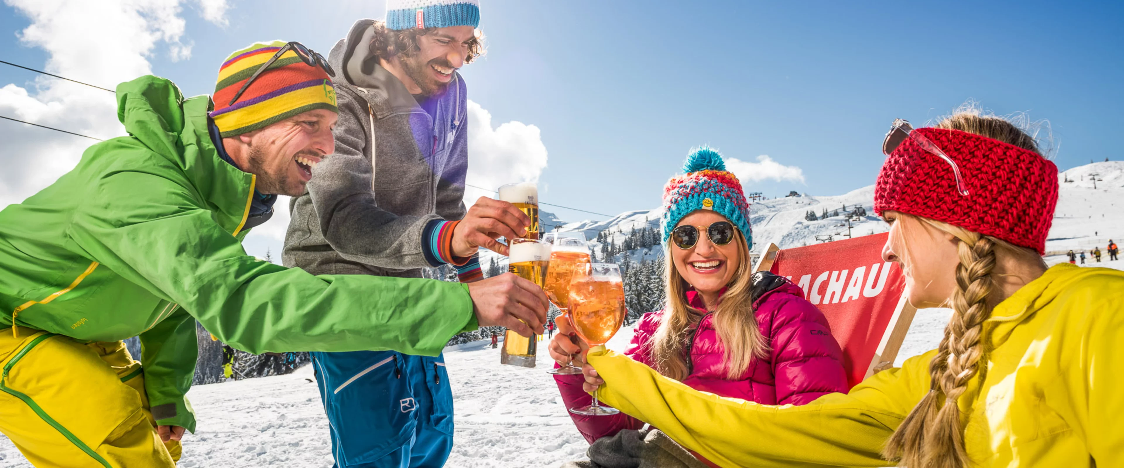 Vier Menschen in farbenfroher Winterkleidung sitzen draußen in einer verschneiten Berglandschaft, stoßen mit Getränken an und lächeln. Sie sind umgeben von schneebedeckten Gipfeln und einem strahlend blauen Himmel.