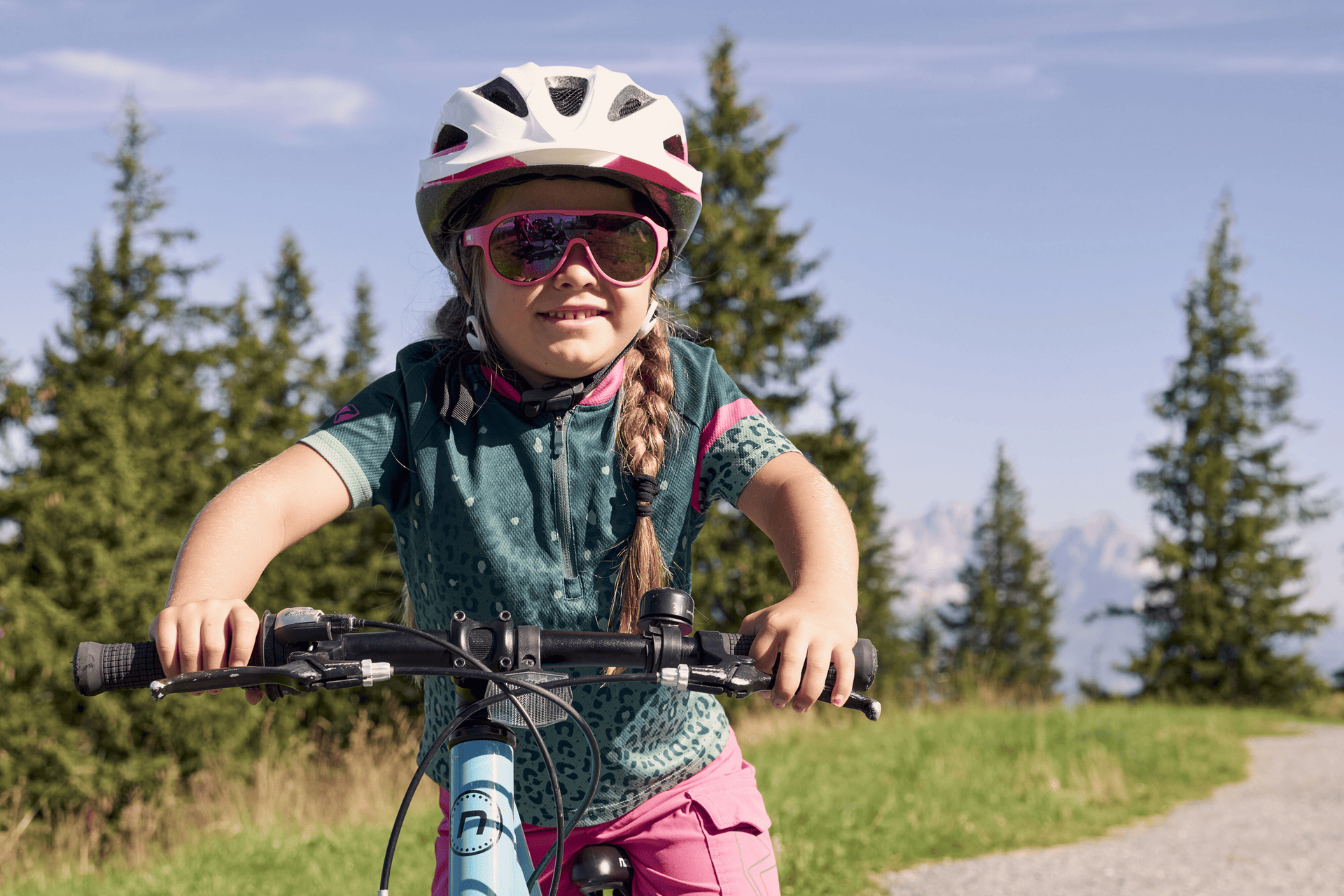 Ein junges Mädchen mit Helm und Sonnenbrille fährt mit dem Fahrrad auf einem Weg, der von grünem Gras und hohen Kiefern umgeben ist, im Hintergrund ein klarer blauer Himmel.