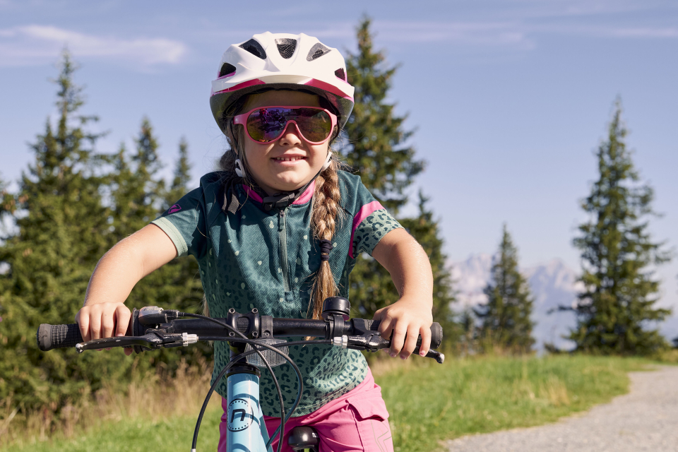 Ein junges Mädchen mit Helm und Sonnenbrille fährt mit dem Fahrrad auf einem Weg, der von grünem Gras und hohen Kiefern umgeben ist, im Hintergrund ein klarer blauer Himmel.