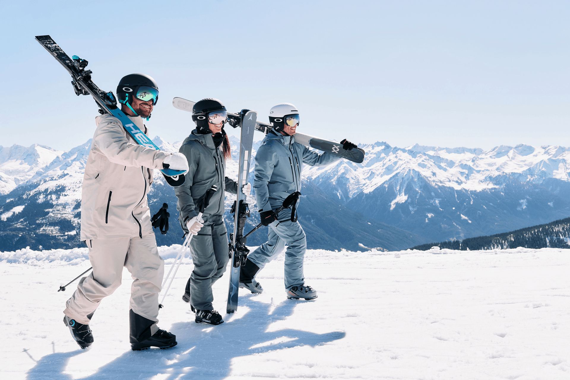 Drei Personen in Skiausrüstung laufen durch eine verschneite Landschaft. Jeder trägt Skier auf seinen Schultern. Sie tragen Winterkleidung und Helme. Im Hintergrund sind schneebedeckte Berge unter einem klaren blauen Himmel zu sehen.