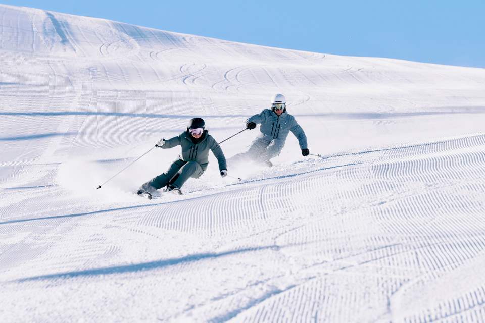 Zwei Skifahrer in Winterkleidung fahren unter einem klaren blauen Himmel eine präparierte Schneepiste hinunter und hinterlassen dabei Spuren im Schnee.