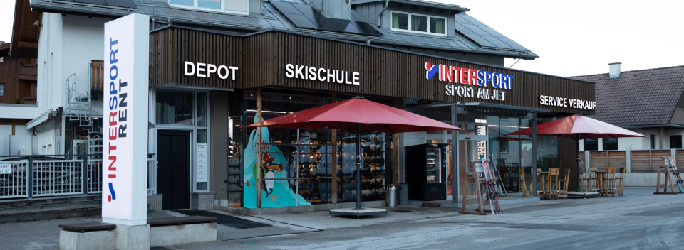 Ein Ladenlokal mit den Schildern „Intersport Rent“, „Depot“, „Skischule“ und „Service Verleih“. Das Gebäude hat eine Holzfassade mit roten Markisen und stellt im Inneren Skiausrüstung aus.