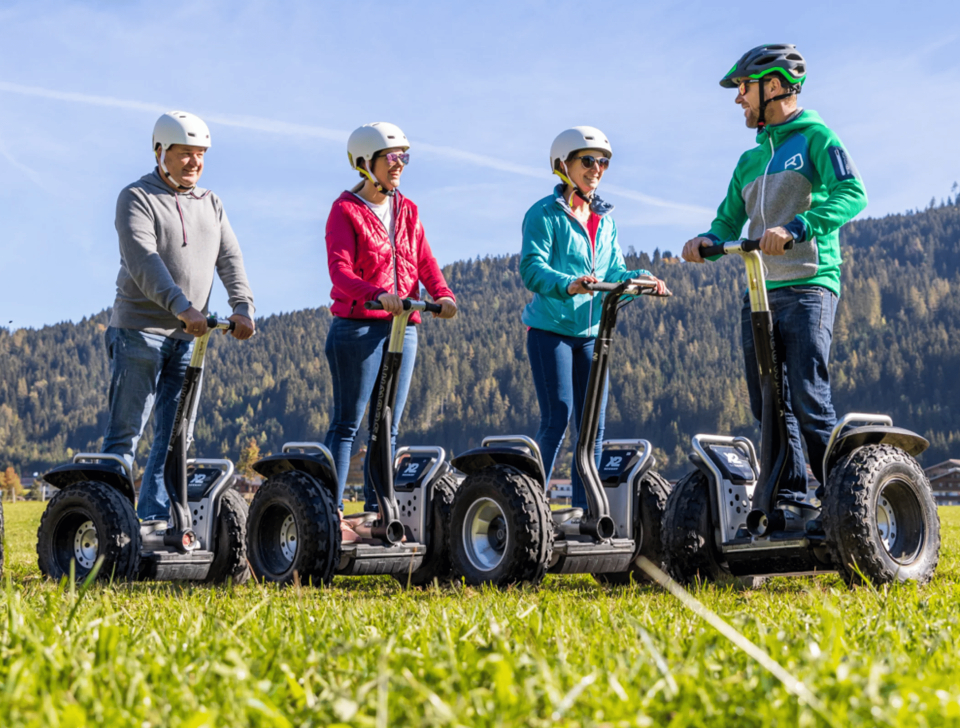 Vier Personen mit Helmen und Freizeitkleidung fahren Segways auf einer Wiese vor der Kulisse bewaldeter Hügel und eines klaren blauen Himmels.