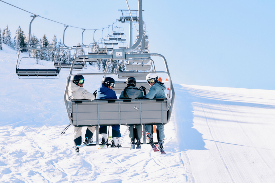Vier Personen in Skiausrüstung sitzen in einem Skilift und fahren einen schneebedeckten Berg hinauf. Der Himmel ist klar und blau, im Hintergrund sind schneebedeckte Bäume zu sehen.