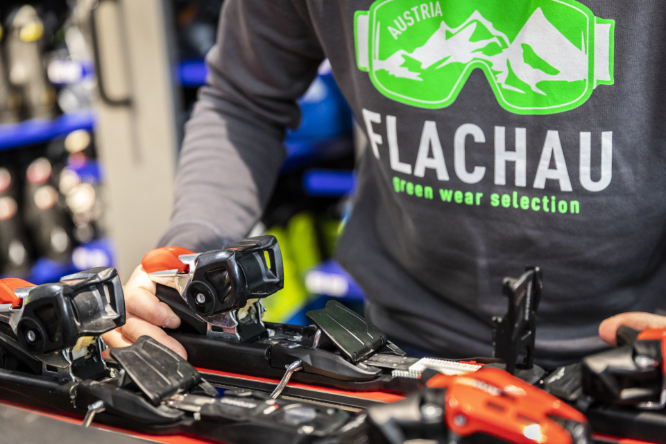 Eine Person in einem grauen Sweatshirt mit grünem Skibrillen-Motiv und dem Schriftzug „Austria Flachau green wear selection“ passt die Skibindung an einem Paar Ski an. Im Hintergrund sind Regale mit Skiausrüstung zu sehen.