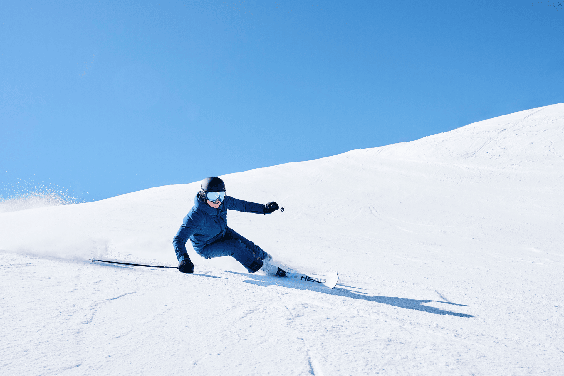 Ein Skifahrer in blauem Outfit und Helm fährt unter einem klaren blauen Himmel eine scharfe Kurve auf einem verschneiten Hang.