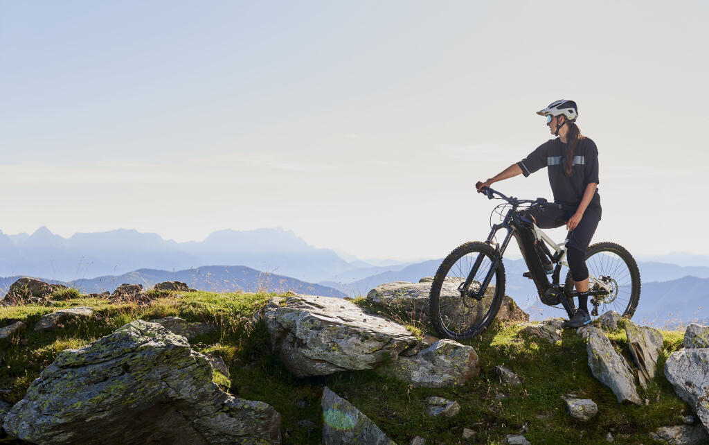 Eine Person mit Helm fährt mit einem Mountainbike auf einem steinigen Pfad mit malerischer Aussicht auf die Berge in der Ferne und einem klaren Himmel im Hintergrund.