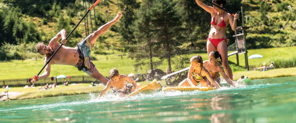 Eine Gruppe von Menschen genießt einen sonnigen Tag an einem See. Eine Person fällt von einem Paddleboard ins Wasser, während andere auf dem Board sitzen und stehen, lachen und planschen. Im Hintergrund sind eine Rasenfläche mit Bäumen und entspannten Menschen zu sehen.