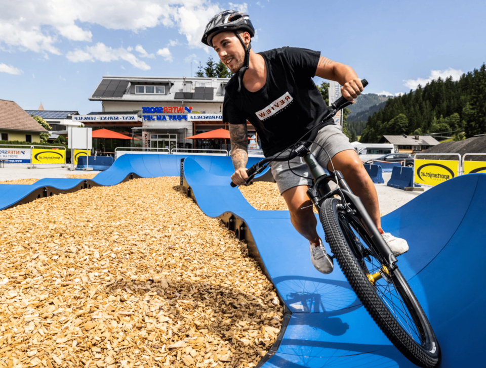 Eine Person mit Helm fährt mit dem Fahrrad auf einem blauen Pumptrack, der mit Holzschnitzeln bedeckt ist. Im Hintergrund sind ein Gebäude mit Schildern und ein bewaldeter Hang unter einem teilweise bewölkten Himmel zu sehen.