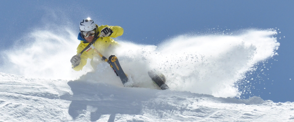 Ein Skifahrer in einer leuchtend gelben Jacke und einem Helm fährt durch den frischen Pulverschnee auf einem verschneiten Hang und erzeugt dabei eine Schneespritze vor einem klaren blauen Himmel.