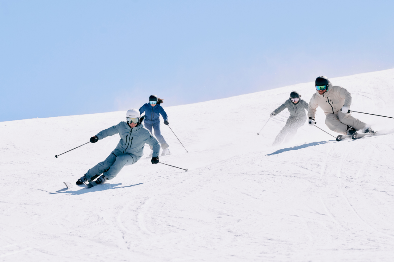 Vier Personen fahren unter einem klaren blauen Himmel einen verschneiten Hang hinunter, jeder trägt Winterkleidung und Helme.