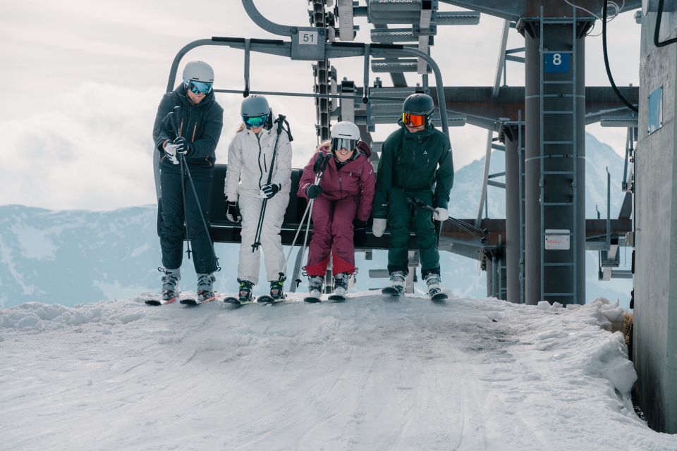 Vier Skifahrer in Winterkleidung sitzen auf einem Skilift am oberen Ende eines schneebedeckten Hangs und bereiten sich auf die Abfahrt vor. Im Hintergrund sind schneebedeckte Berge unter einem bewölkten Himmel zu sehen.